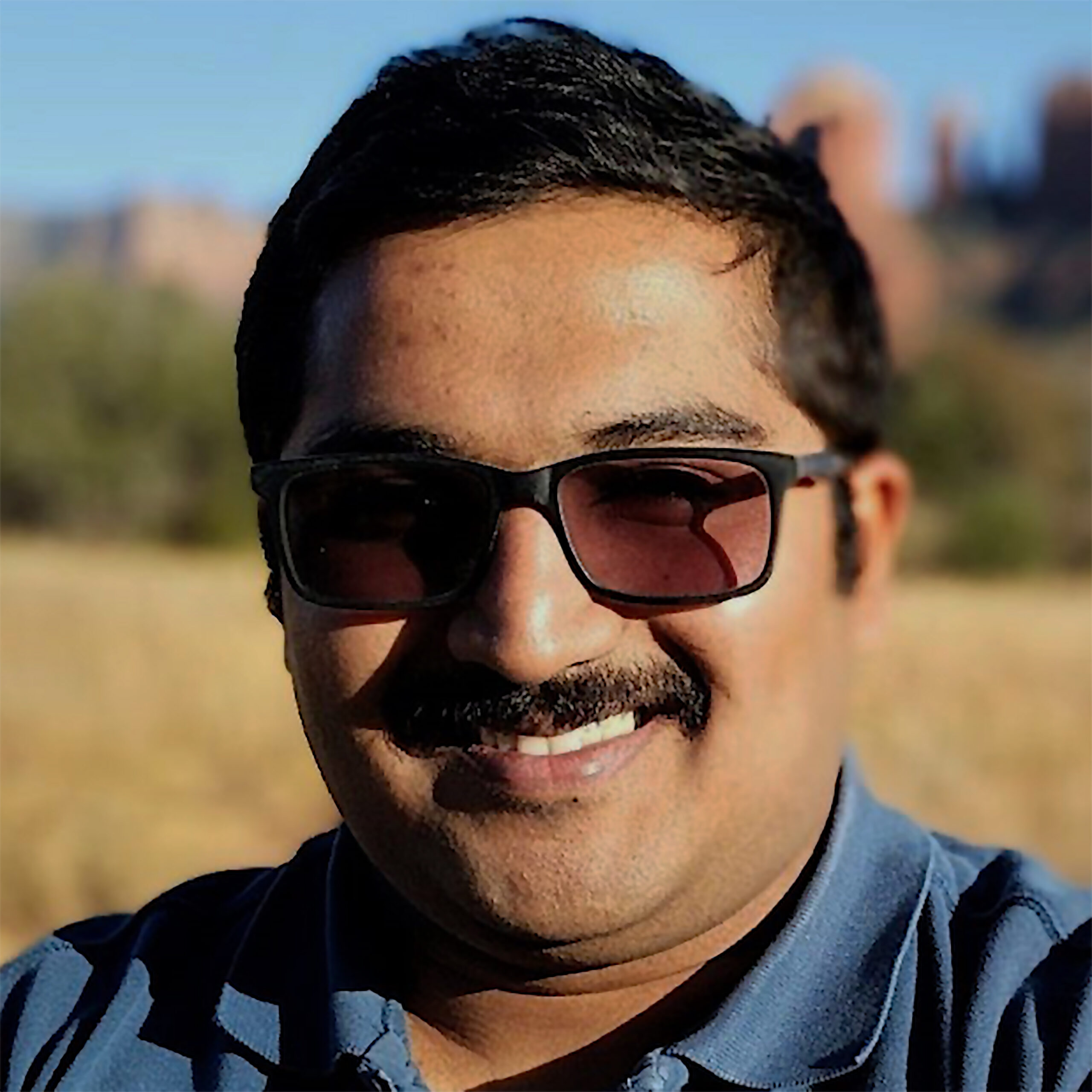 A smiling individual wearing sunglasses, with a blurred natural background.