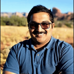 A smiling individual wearing sunglasses, with a blurred natural background.